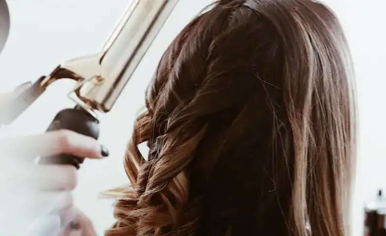 A hairdresser using a curling iron to create waves in a woman's long, brown hair during a professional styling session.