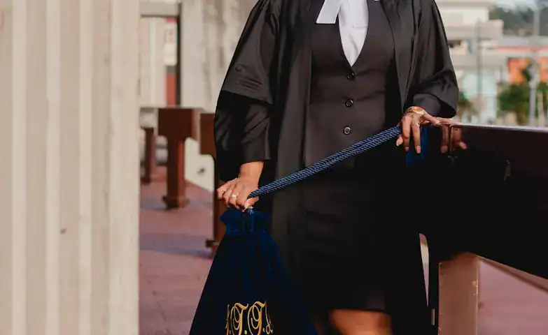 A person in a graduation gown and cap holding a diploma in a court or legal setting, with a wooden bench and urban background.