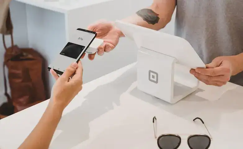 A person making a contactless payment using a smartphone at a checkout counter, with another person holding a receipt or invoice. Sunglasses are placed on the counter, and a small white device is used.