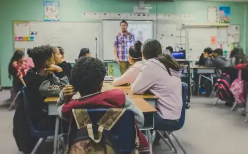 Students attentively listening to teacher in a modern classroom setting with educational posters and technology.