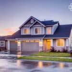 A contemporary two-storey house with a three-car garage, large windows, and a neatly landscaped front yard, set against a colourful sunset sky. The wet driveway reflects the warm lighting from the hou.