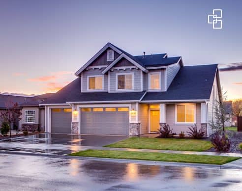 A contemporary two-storey house with a three-car garage, large windows, and a neatly landscaped front yard, set against a colourful sunset sky. The wet driveway reflects the warm lighting from the hou.