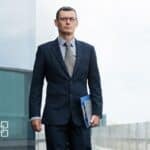 A professional man in a navy suit and glasses standing outdoors, holding a blue folder, with a modern building in the background, representing business and financial services.