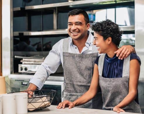 Two cheerful cafe staff members, a man and a woman, smiling and enjoying their work in a contemporary kitchen setting, wearing aprons and engaging with each other.