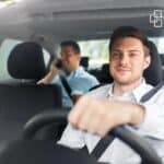 A young man driving a car, with another person in the back seat talking on the phone, representing professional service and client support.
