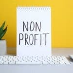 Notepad displaying "Non Profit" on a desk with a keyboard, mouse, and potted plant, set against a yellow background, representing non-profit organisation themes.