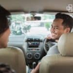 Smiling man driving a car while talking to a passenger, enjoying a relaxed moment on a sunny day.