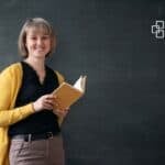 Young woman smiling with a book in front of a blackboard at school or university.