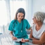A caring nurse in scrubs provides medical support and comfort to an elderly woman during a consultation in a bright, modern healthcare setting.