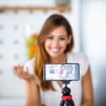 Smiling woman in a white top recording a video with her smartphone mounted on a tripod, indoors with a blurred background.