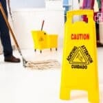 Bright yellow caution sign warning of a wet floor in a commercial or retail environment, with mop and cleaning supplies visible in the background.