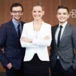 Three confident professionals, two men and one woman, dressed in suits, standing together with a warm smile.