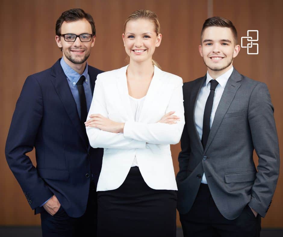 Three confident professionals, two men and one woman, dressed in suits, standing together with a warm smile.