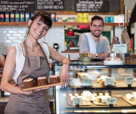 Smiling female barista holding a tray of assorted cakes in a cozy café setting with a male colleague in the background.