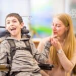 A young boy with a disability in a wheelchair smiling and engaging with a woman, possibly a caregiver or teacher, in a supportive learning environment.