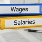 Understanding the Criminalisation of Wage Theft Close-up of blue and yellow filing binders labeled "Wages" and "Salaries" on a desk with a pen and open documents, representing payroll and employee compensation management.
