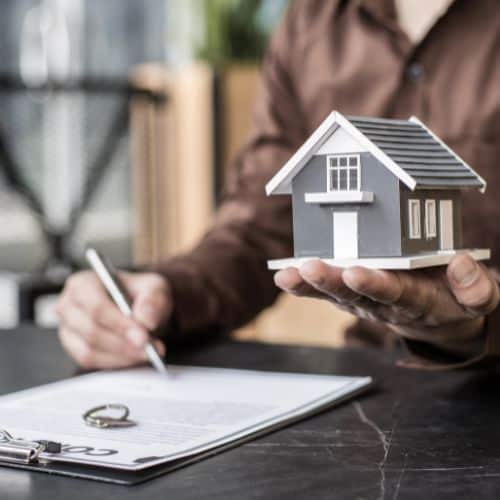 House model for real estate or property investment, held by a person with a pen and documents on a table.