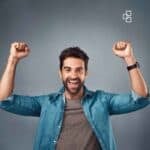 How to Choose a Top Small Business Accountant for Your Growth A cheerful man with dark hair and a beard, wearing a denim shirt and grey t-shirt, smiling broadly with arms raised in celebration against a plain grey background.