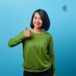 A cheerful woman with shoulder-length black hair, wearing a green sweater, giving a thumbs-up gesture against a blue background, representing positive client service and trust.