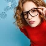 A young woman with curly red hair, wearing black glasses and a red sweater, looks directly at the camera while adjusting her glasses, with a background featuring digital fingerprint and DNA icons.