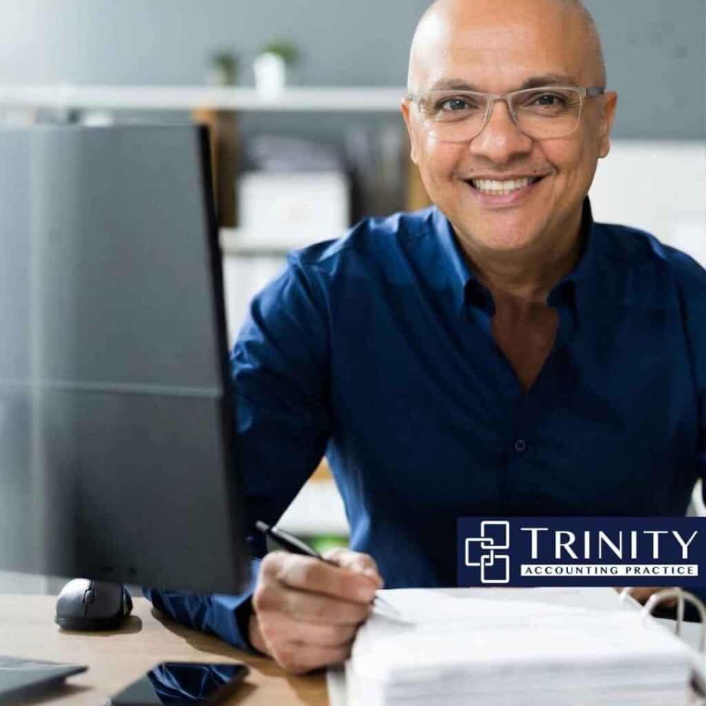 Smiling man in blue shirt working at desk with computer, representing Trinity Accounting Practice's professional tax and accounting services in Beverly Hills, NSW.