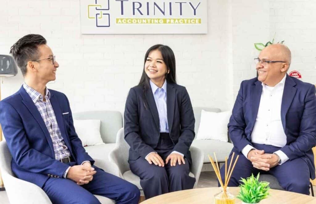 Three professionals in business attire having a discussion in an office with Trinity Accounting Practice sign.