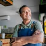 Small Business Accounting Guide for Australian SMEs A cheerful male accountant with crossed arms, wearing a green shirt and blue apron, standing confidently in a professional workspace.