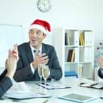 Holiday Cash Flow Survival Guide for Small Business Business professionals celebrating during a holiday meeting, with one person wearing a Santa hat, in a modern office setting with documents and glasses of champagne.