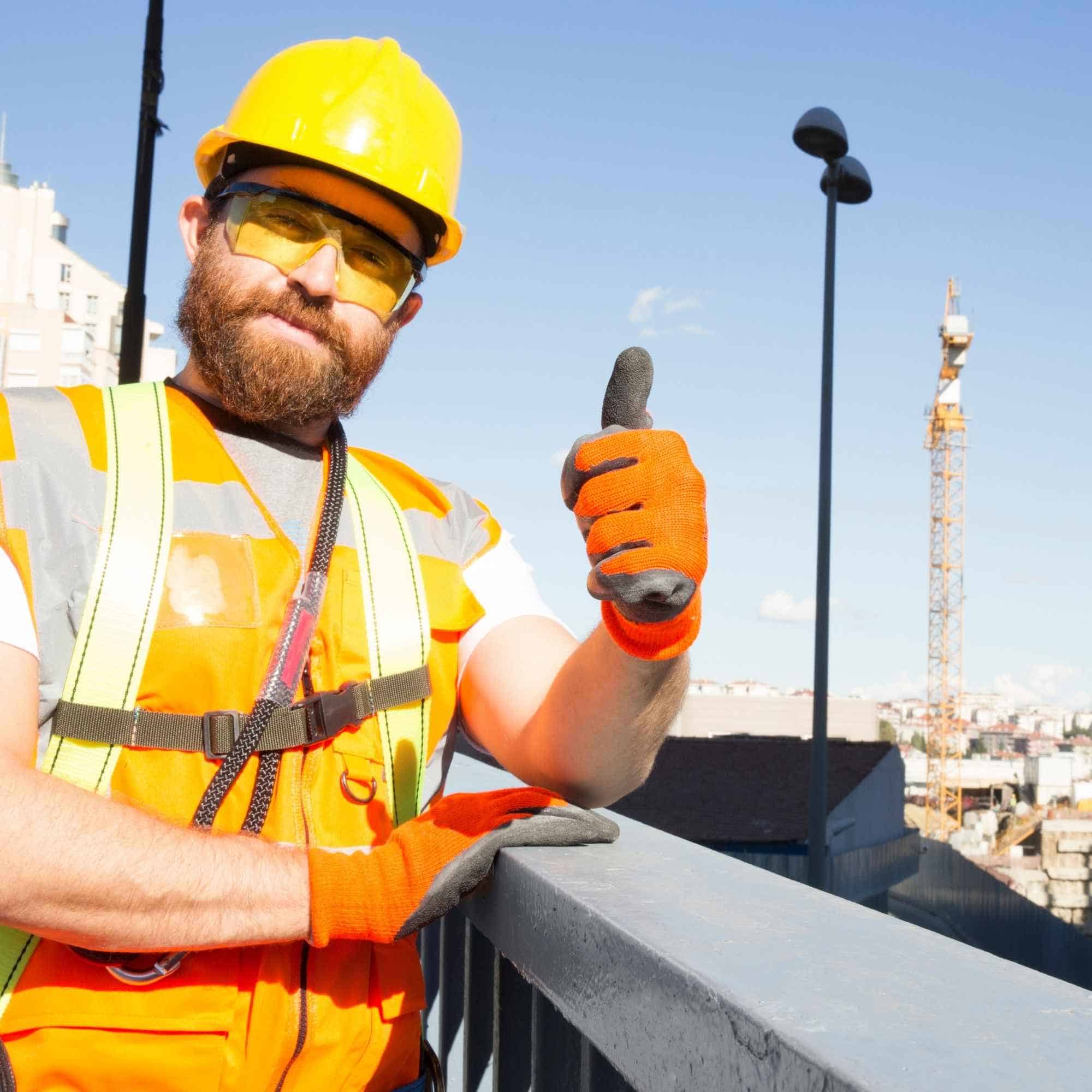 Construction worker in safety gear, including a yellow helmet and orange gloves, standing on a rooftop with a cityscape and crane in the background.