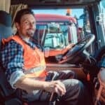 A truck driver in a high-visibility vest sitting in the cab of a large truck, smiling at the camera with a view of the road and other trucks outside.