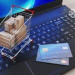 Small shopping cart filled with cardboard boxes placed on a laptop keyboard alongside a credit card, symbolising online shopping, e-commerce, or financial transactions.