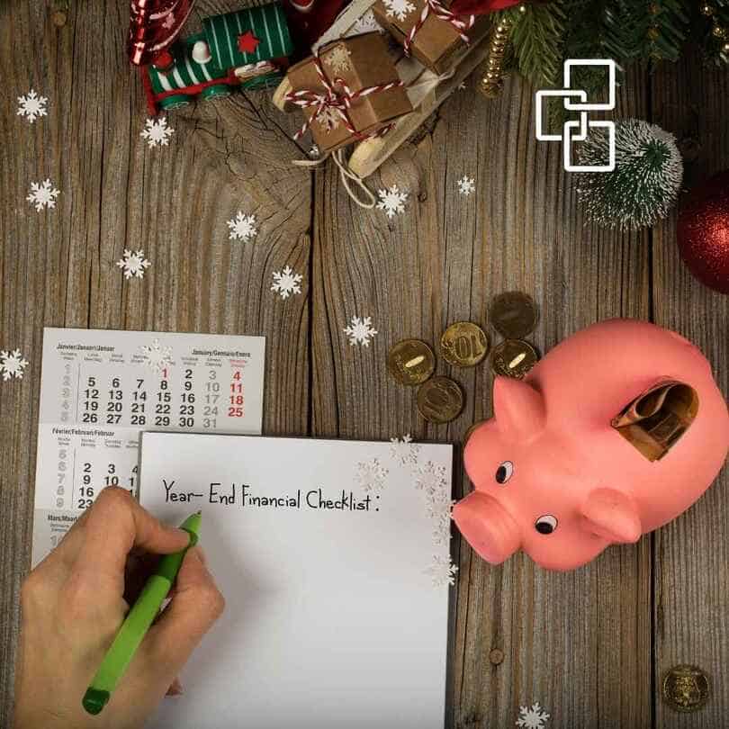Hand writing "Year-End Financial Checklist" on paper with a pink piggy bank, coins, and Christmas decorations on a wooden table, symbolising financial review and savings during the festive season.