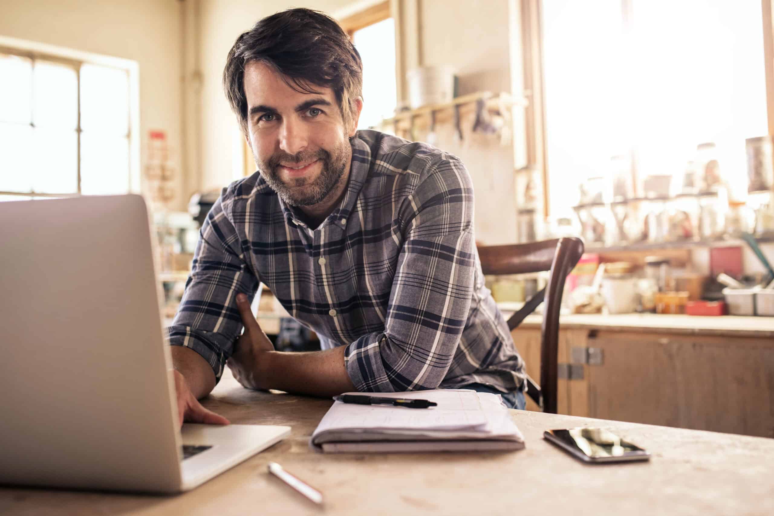 A smiling man with dark hair and a beard working on a laptop at a desk in a bright, well-lit office with large windows and shelves in the background.