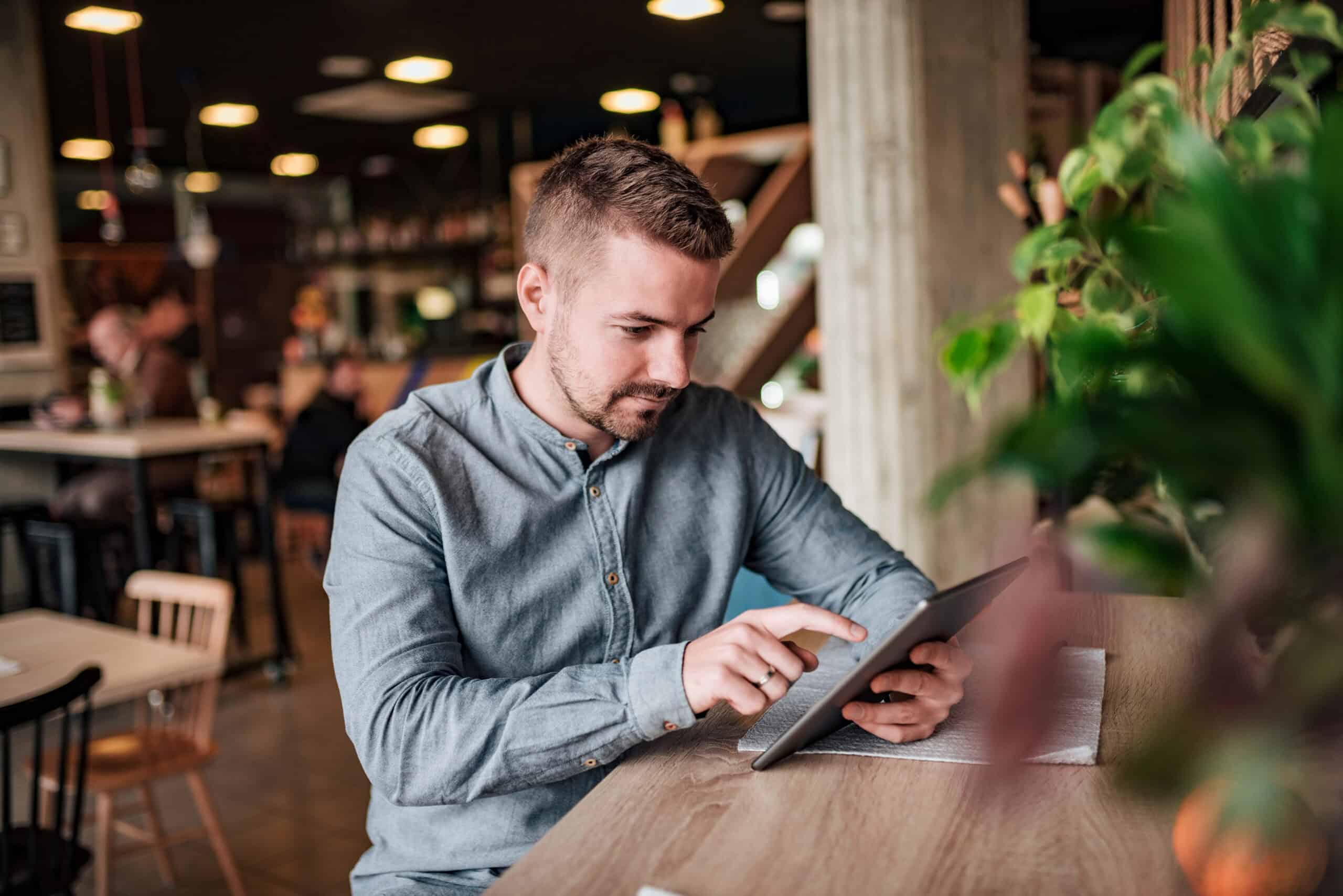 Young man in casual attire working on a tablet at a café, with a focus on digital technology and modern workspace environment.