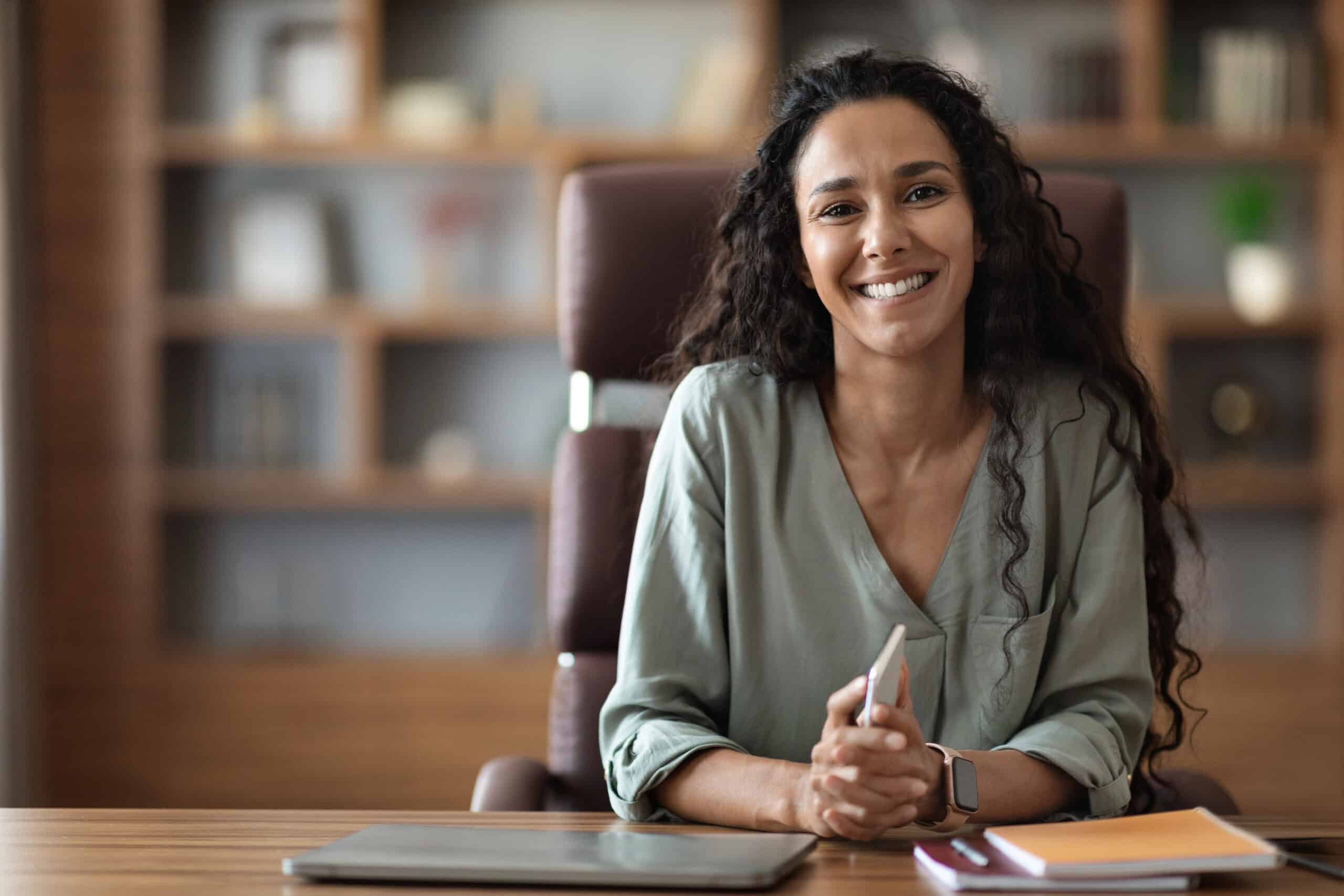 A smiling woman with curly hair sitting at a desk, holding a smartphone, in a professional office environment.