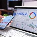 How Does Your Business Compare? Try Our New Industry Benchmark Tool A person holding a smartphone displaying financial charts, with a laptop screen in the background showing various data visualisations for accounting and business analysis.