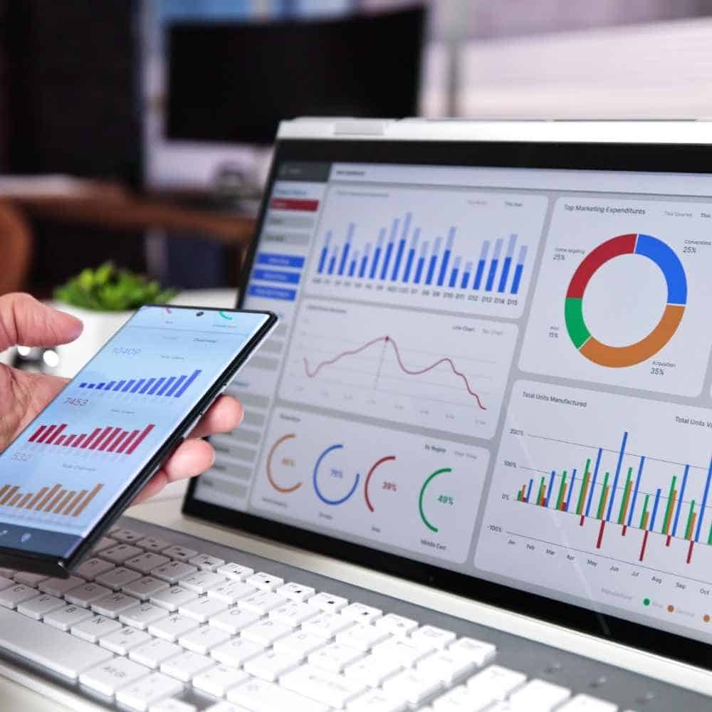 A person holding a smartphone displaying financial charts, with a laptop screen in the background showing various data visualisations for accounting and business analysis.