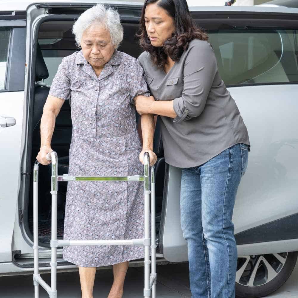 Elderly woman using a walker being assisted by a caregiver outside a vehicle, demonstrating support and care for seniors.